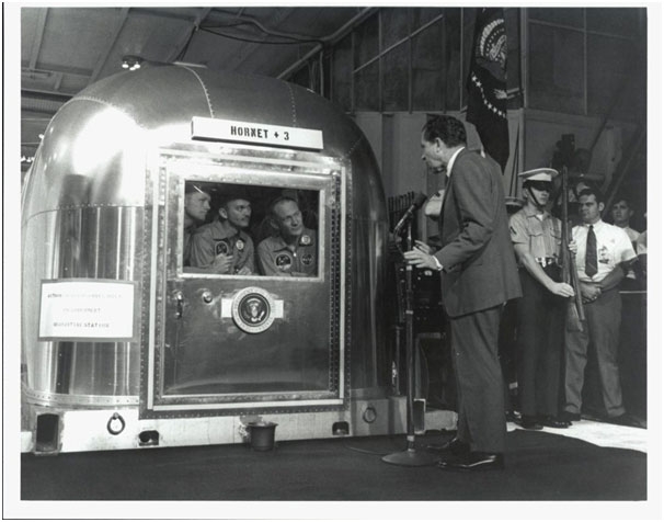 The Apollo 11 astronauts, left to right, Neil A. Armstrong, Michael Collins & Edwin E. Aldrin Jr., inside the Mobile Quarantine Facility aboard the USS Hornet, listen to President Richard M. Nixon.